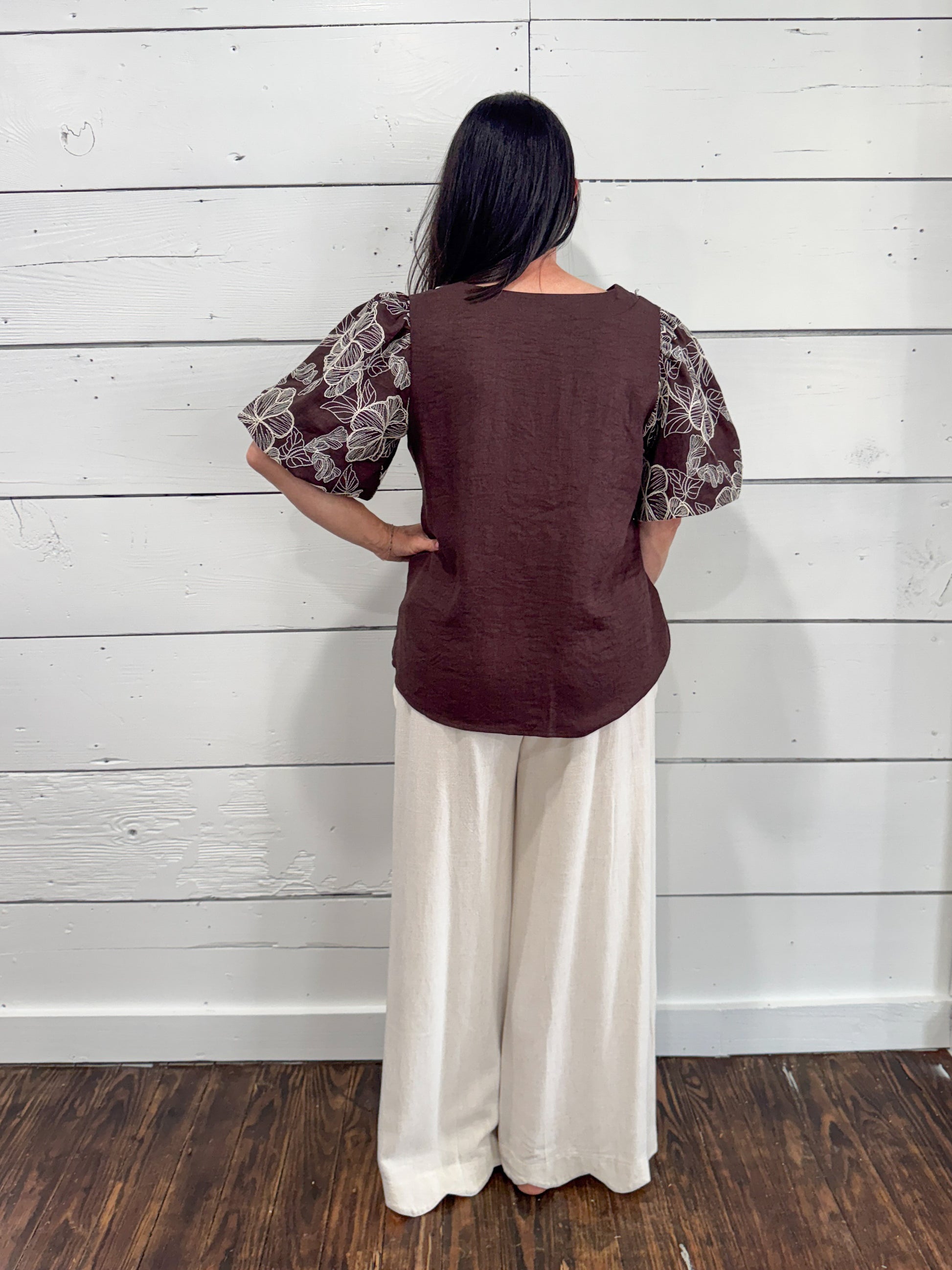 A woman shown from the back wearing a brown linen vest with patterned sheer puff sleeves and cream wide-leg pants, standing against a white wood-paneled wall.