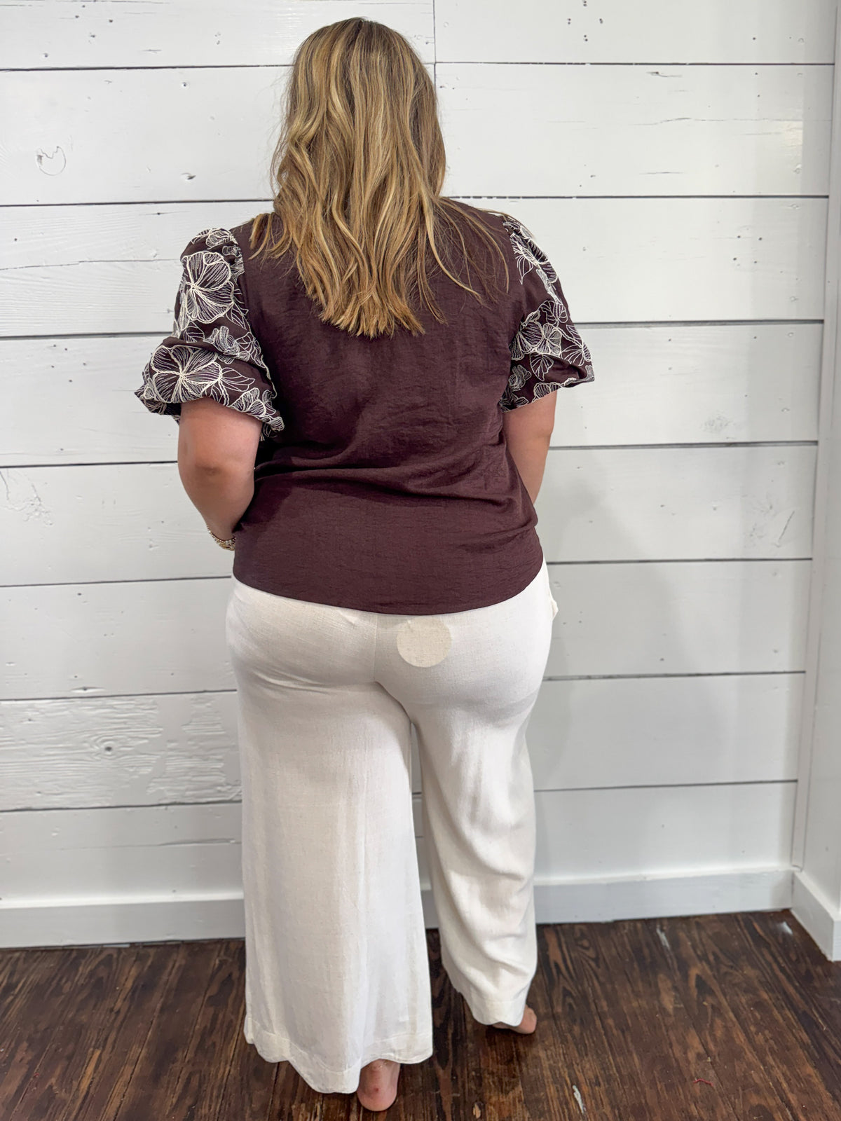 A woman viewed from behind wearing a dark brown sleeveless top with patterned short puff sleeves and white wide-leg pants, standing against a white shiplap wall on a dark wood floor.