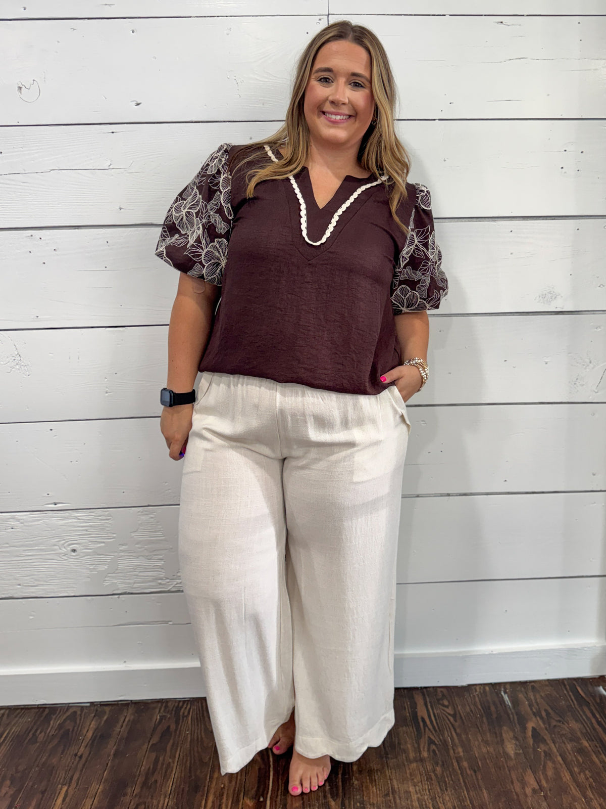 A woman modeling a dark brown short-sleeve top with patterned sheer puff sleeves and cream wide-leg pants, standing against a white wood-paneled wall.