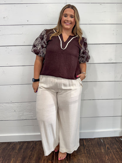 A woman modeling a dark brown short-sleeve top with patterned sheer puff sleeves and cream wide-leg pants, standing against a white wood-paneled wall.