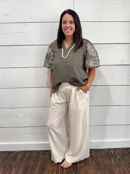 A woman stands barefoot against a white wood plank wall, wearing a loose olive green top with sheer floral-sleeve sleeves and cream wide-leg pants.