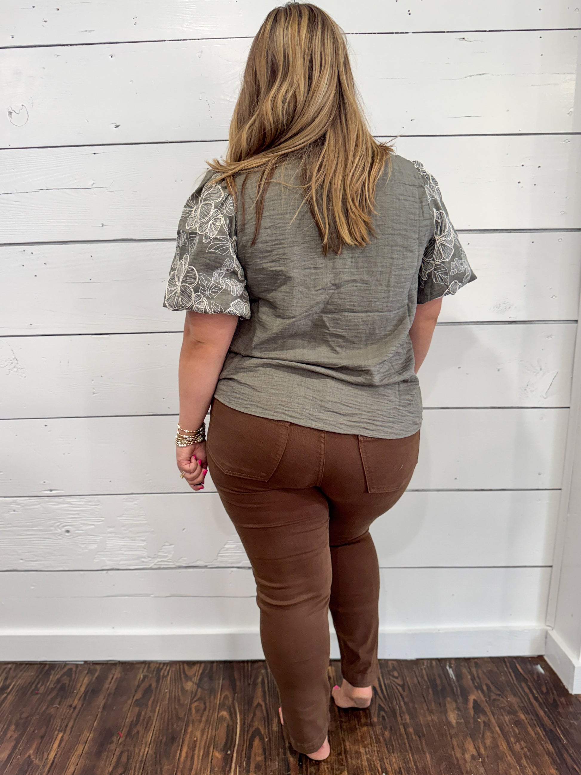 A woman viewed from behind wearing a gray short-sleeve top with patterned sleeves and brown pants, standing against a white shiplap wall on a dark wood floor.