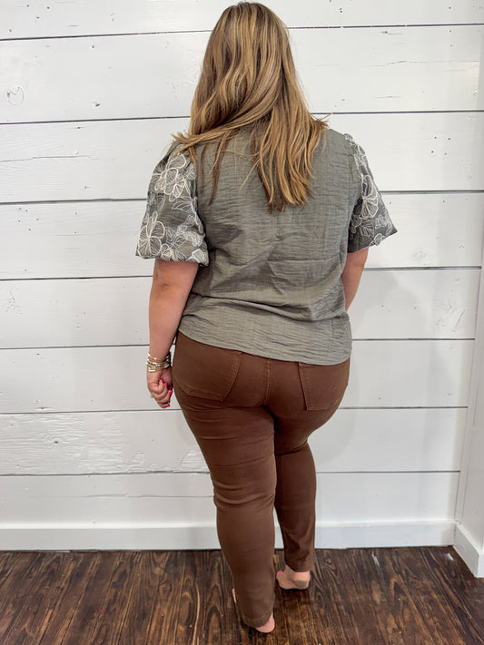 A woman viewed from behind wearing a gray short-sleeve top with patterned sleeves and brown pants, standing against a white shiplap wall on a dark wood floor.