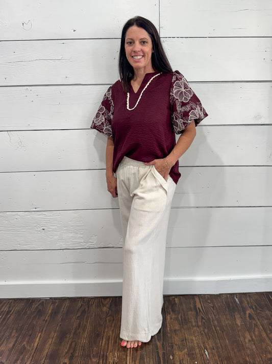 A woman modeling a burgundy short-sleeve top with sheer floral-patterned sleeves and cream wide-leg pants, standing against a white wood-paneled wall.