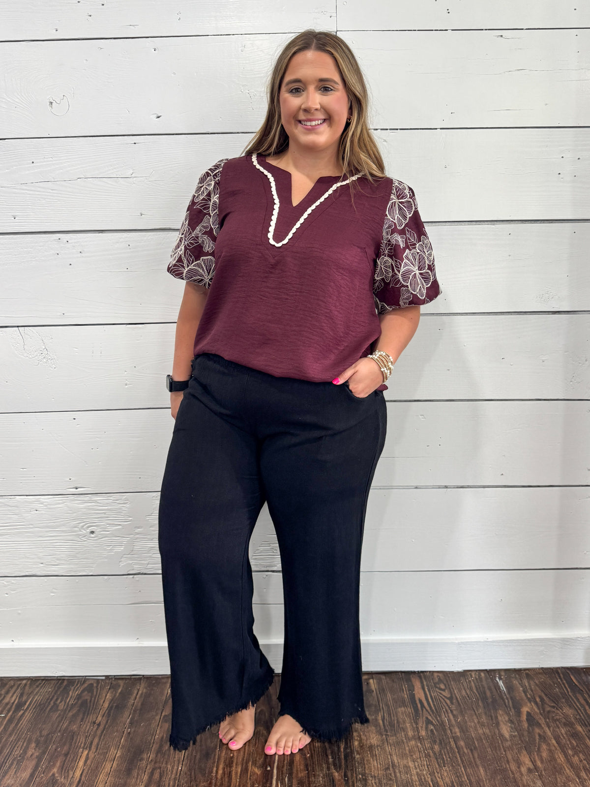 A woman modeling a two-piece outfit consisting of a maroon short-sleeve top with white lace sleeves and a white beaded collar, paired with black wide-leg pants, standing against a white wood-paneled wall.