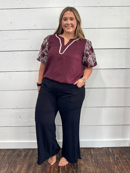 A woman modeling a two-piece outfit consisting of a maroon short-sleeve top with white lace sleeves and a white beaded collar, paired with black wide-leg pants, standing against a white wood-paneled wall.