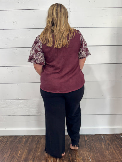 A woman viewed from behind wearing a burgundy top with patterned short sleeves and black pants, standing against a white wood-paneled wall on a dark wood floor.