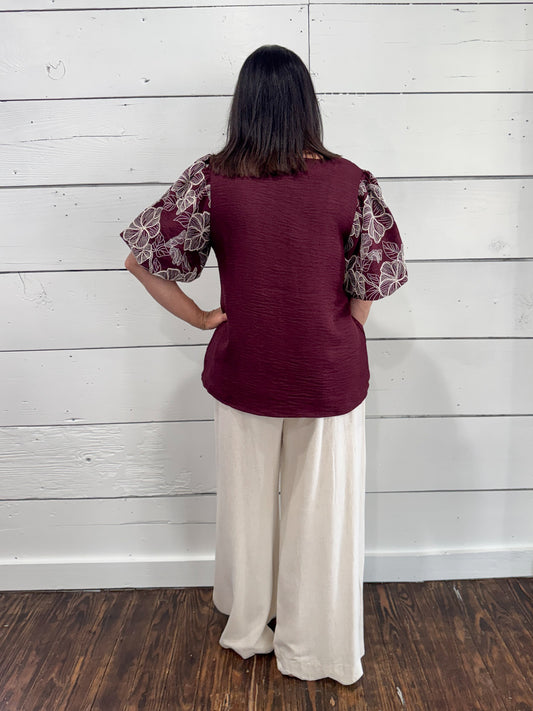 A woman viewed from behind wearing a textured deep burgundy top with patterned flutter sleeves and cream wide-leg pants, standing against a white wood-paneled wall.