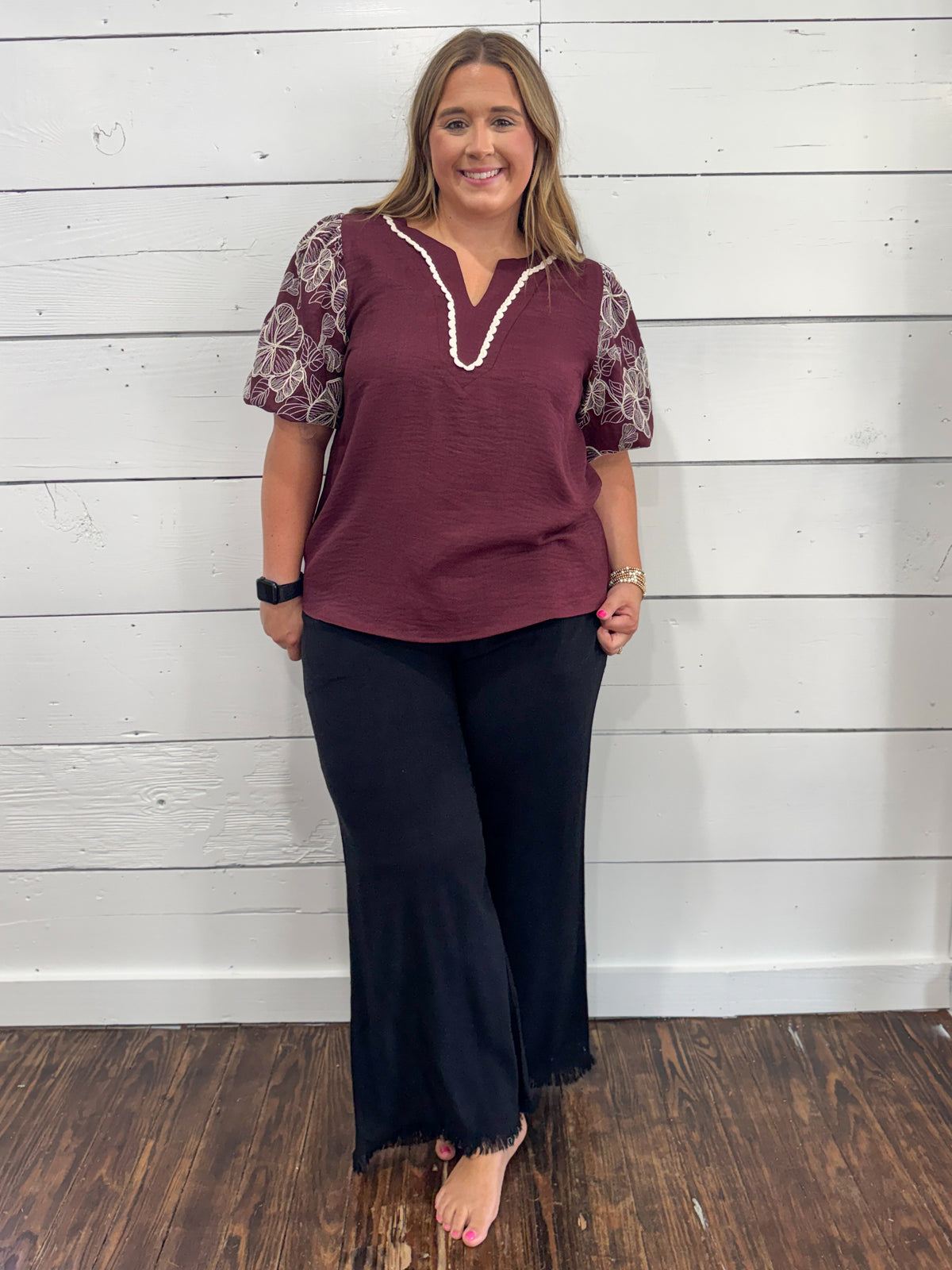 A woman modeling a short-sleeve top with a burgundy body, white lace trim at the split, and patterned white sleeves, paired with dark pants, standing against a white wood-paneled wall.
