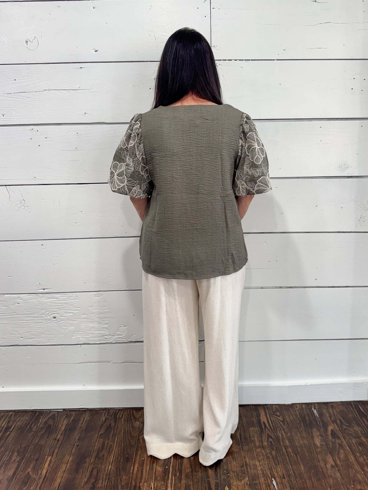 A woman viewed from the back wearing a textured sage green top with patterned puff sleeves and cream wide-leg pants, standing against a white wood plank wall on a dark wood floor.