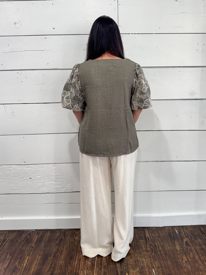 A woman viewed from the back wearing a textured sage green top with patterned puff sleeves and cream wide-leg pants, standing against a white wood plank wall on a dark wood floor.