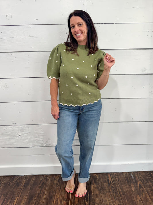 A woman modeling a sage green short-sleeve knit top with white polka dots and scalloped edges, paired with blue jeans, standing against a white wood-paneled wall.