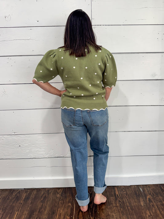 A woman viewed from behind wearing an olive green knit sweater with white polka dots and puff sleeves, paired with blue jeans, standing against a white wood-paneled wall.