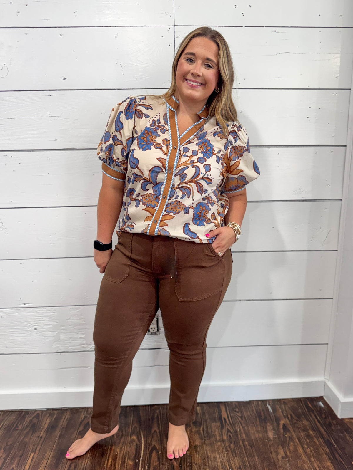 A woman modeling a short-sleeve button-front blouse with a beige, brown, and blue floral pattern, paired with brown pants, standing against a white shiplap wall.