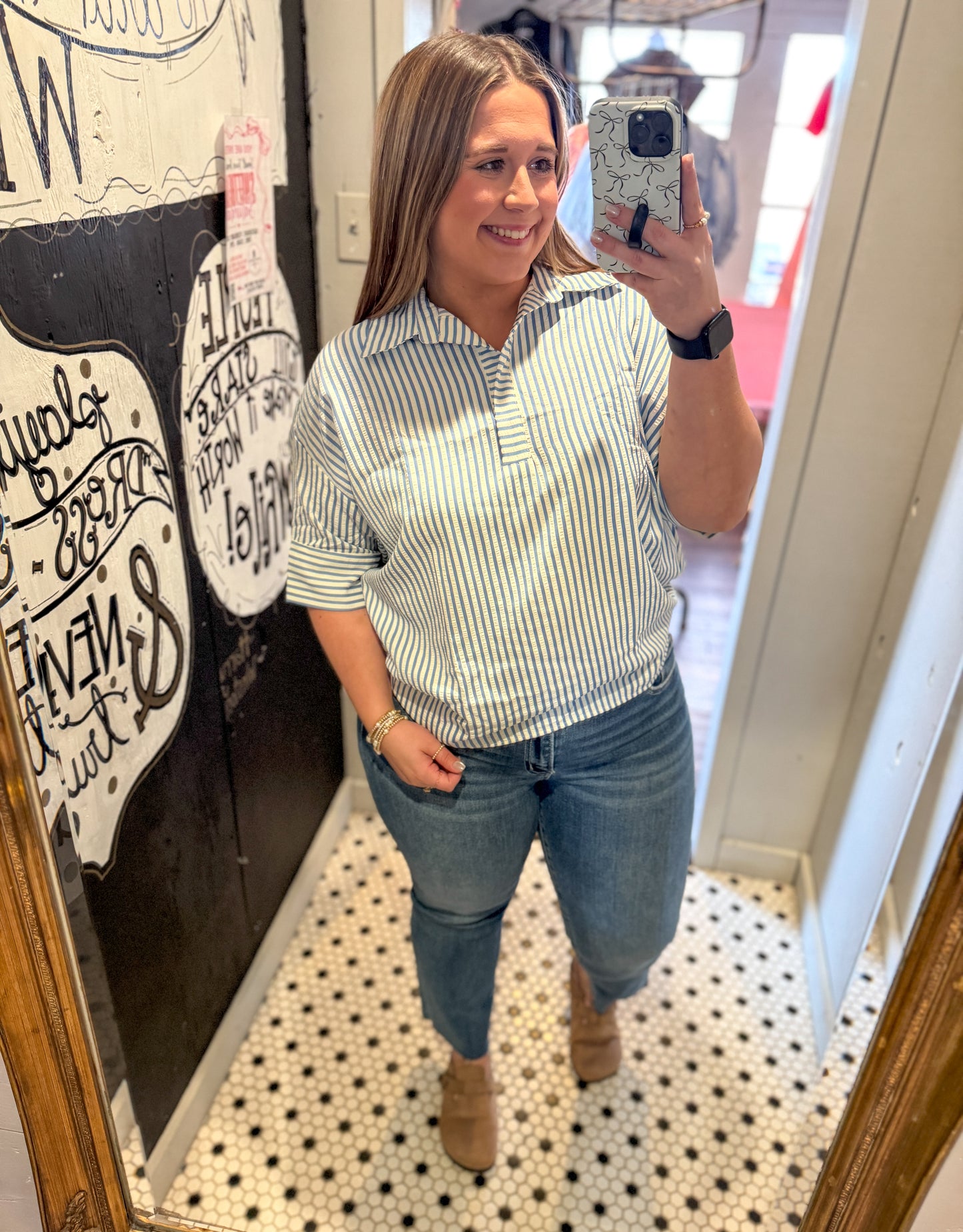 Woman taking a mirror selfie wearing a striped shirt and jeans in a room with decorative elements.