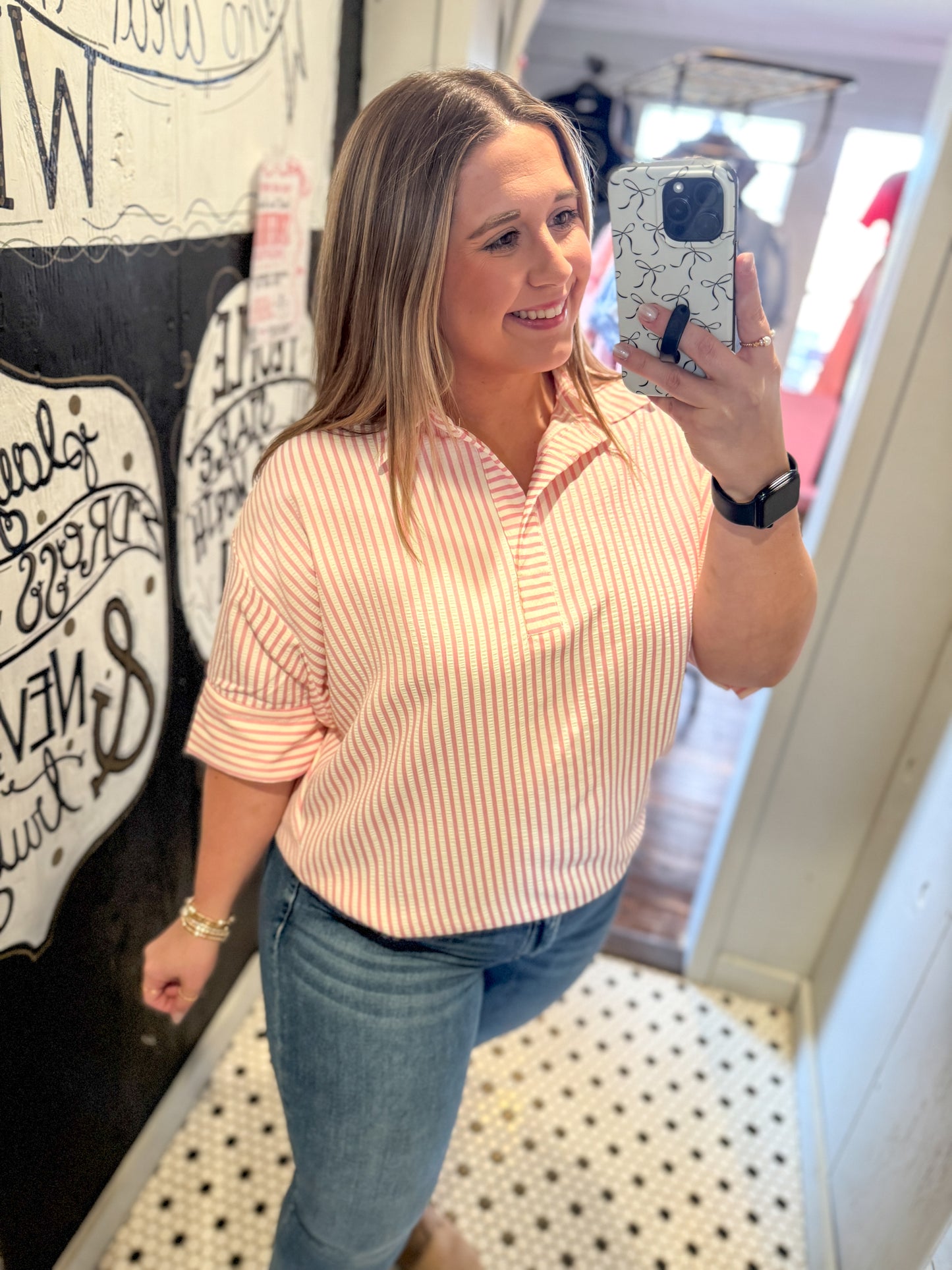 Woman taking a mirror selfie wearing a pink striped shirt and blue jeans in a store.