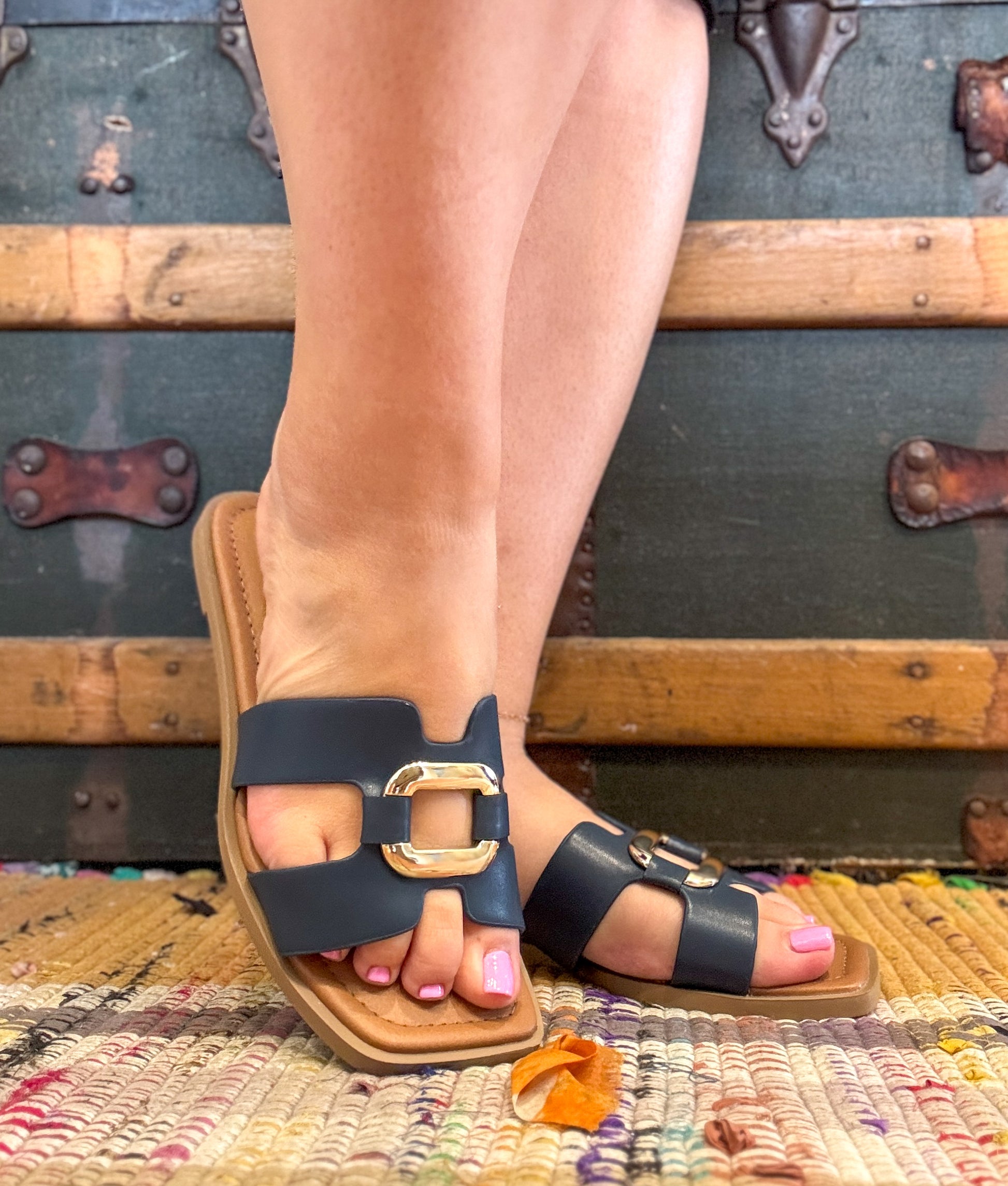 Person wearing navy sandals with gold buckles on a textured rug with vintage suitcases in the background.