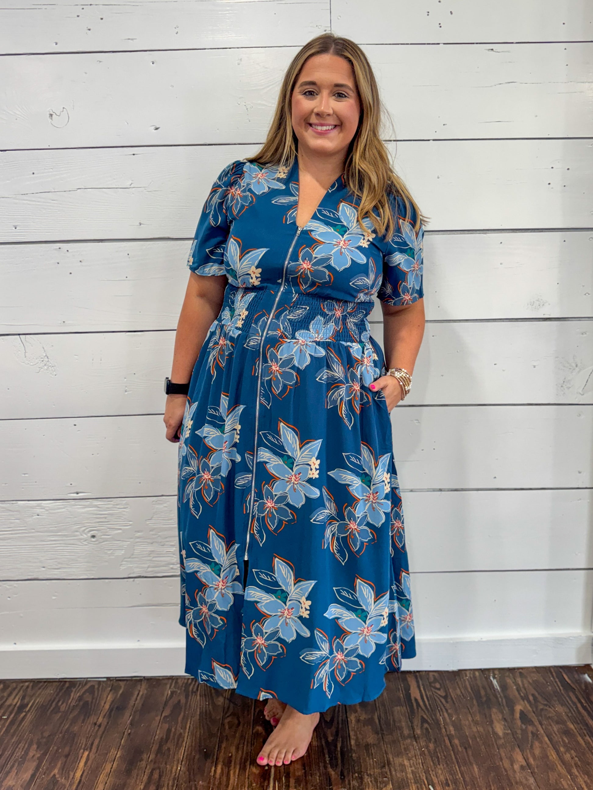 A woman modeling a blue midi wrap dress with a white and light blue floral pattern, standing against a white wood-paneled wall.