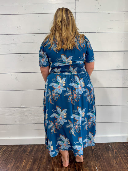 A woman viewed from behind wearing a blue midi dress with a light blue, white, and orange floral pattern, standing against a white wood-paneled wall on a dark wood floor.