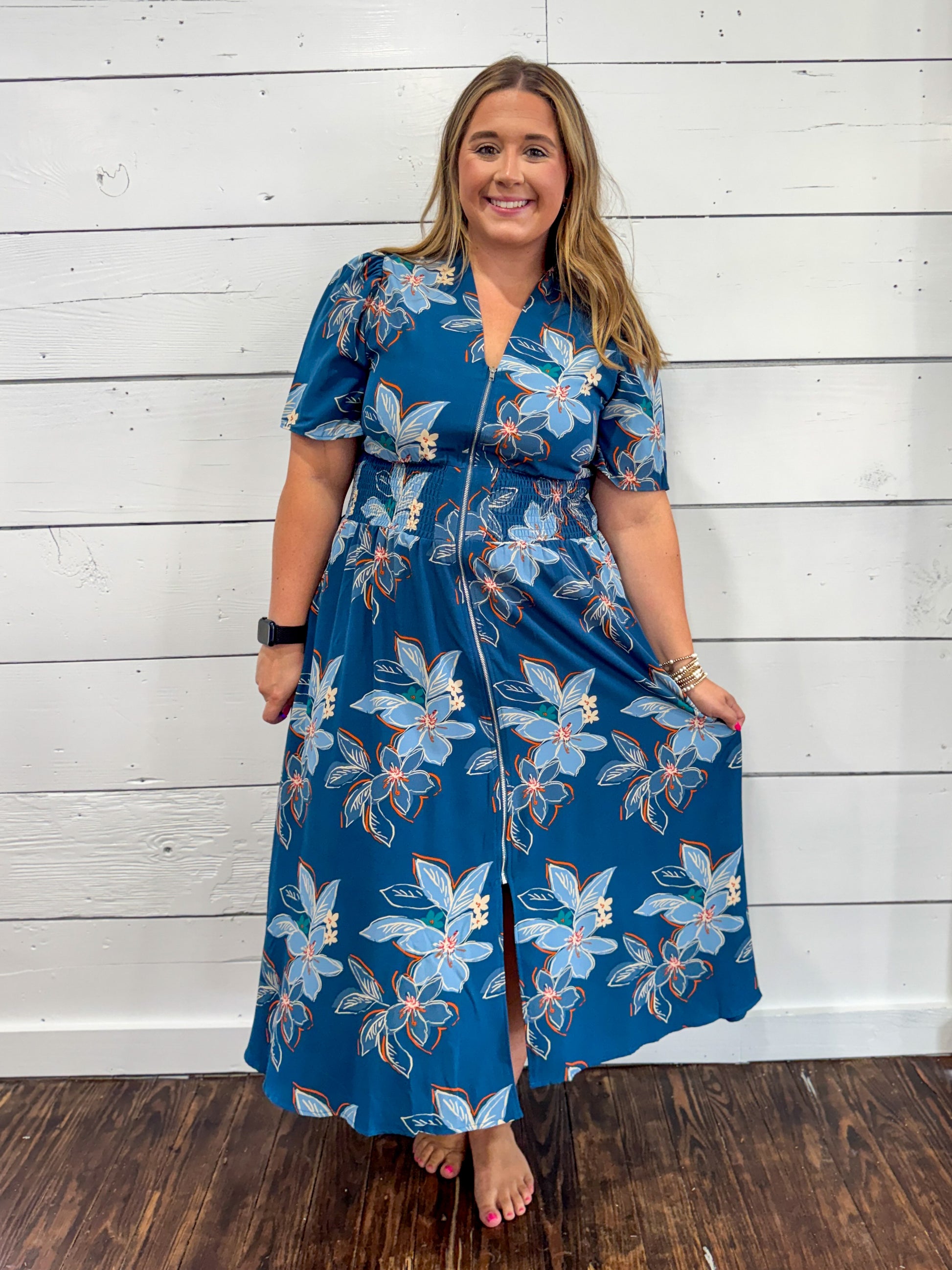 A woman modeling a blue floral midi dress with puff sleeves, a split neckline, and a front slit, standing against a white wood plank wall on a dark wood floor.