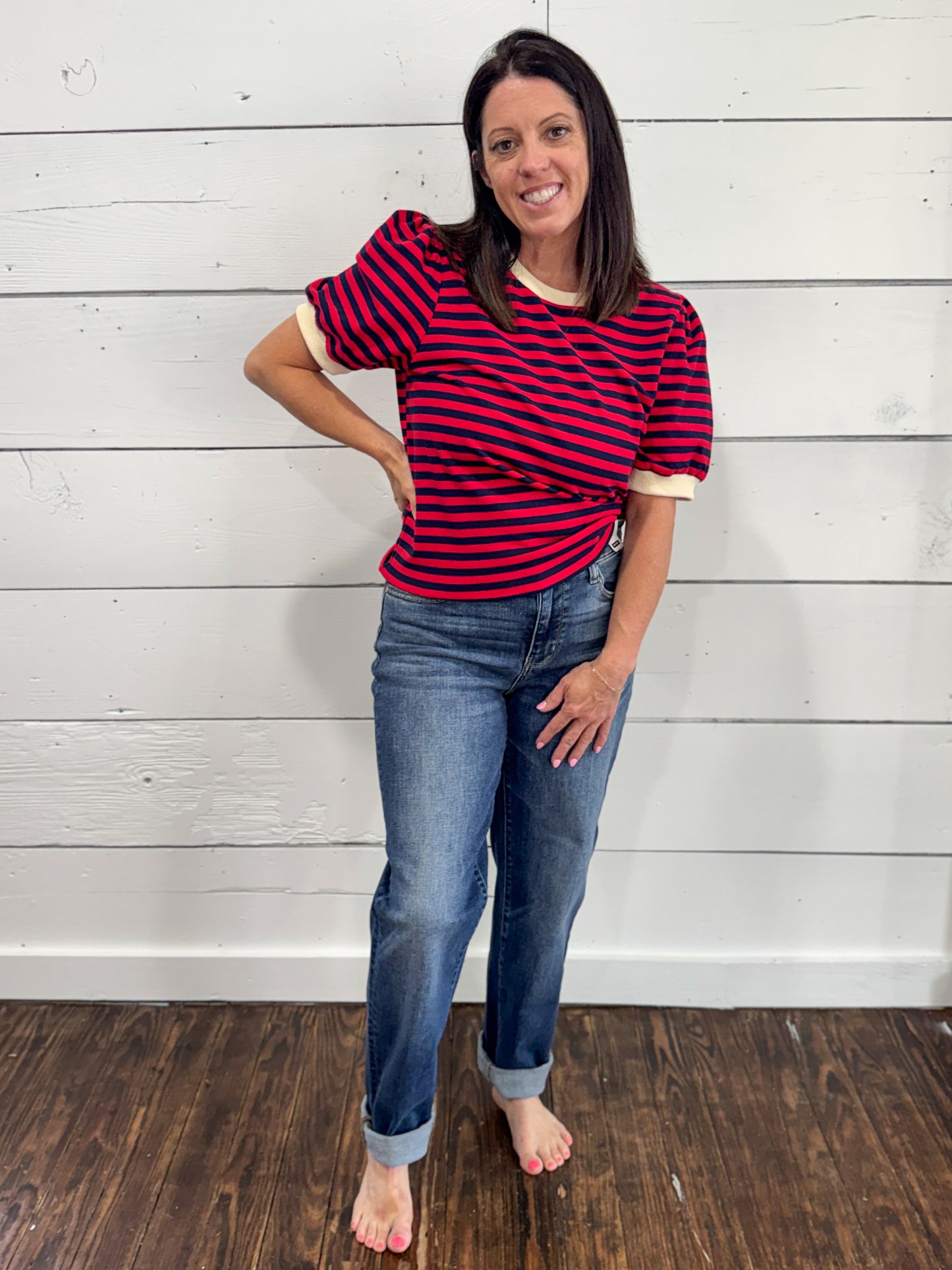 A woman modeling a short-sleeve t-shirt with red and navy horizontal stripes and cream ribbed trim, paired with blue jeans, standing against a white wood-paneled wall.