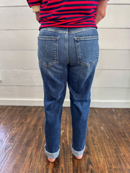 Rear view of a person wearing blue denim jeans with rolled cuffs and a red-and-navy striped shirt, standing on a wood floor against a white paneled wall.