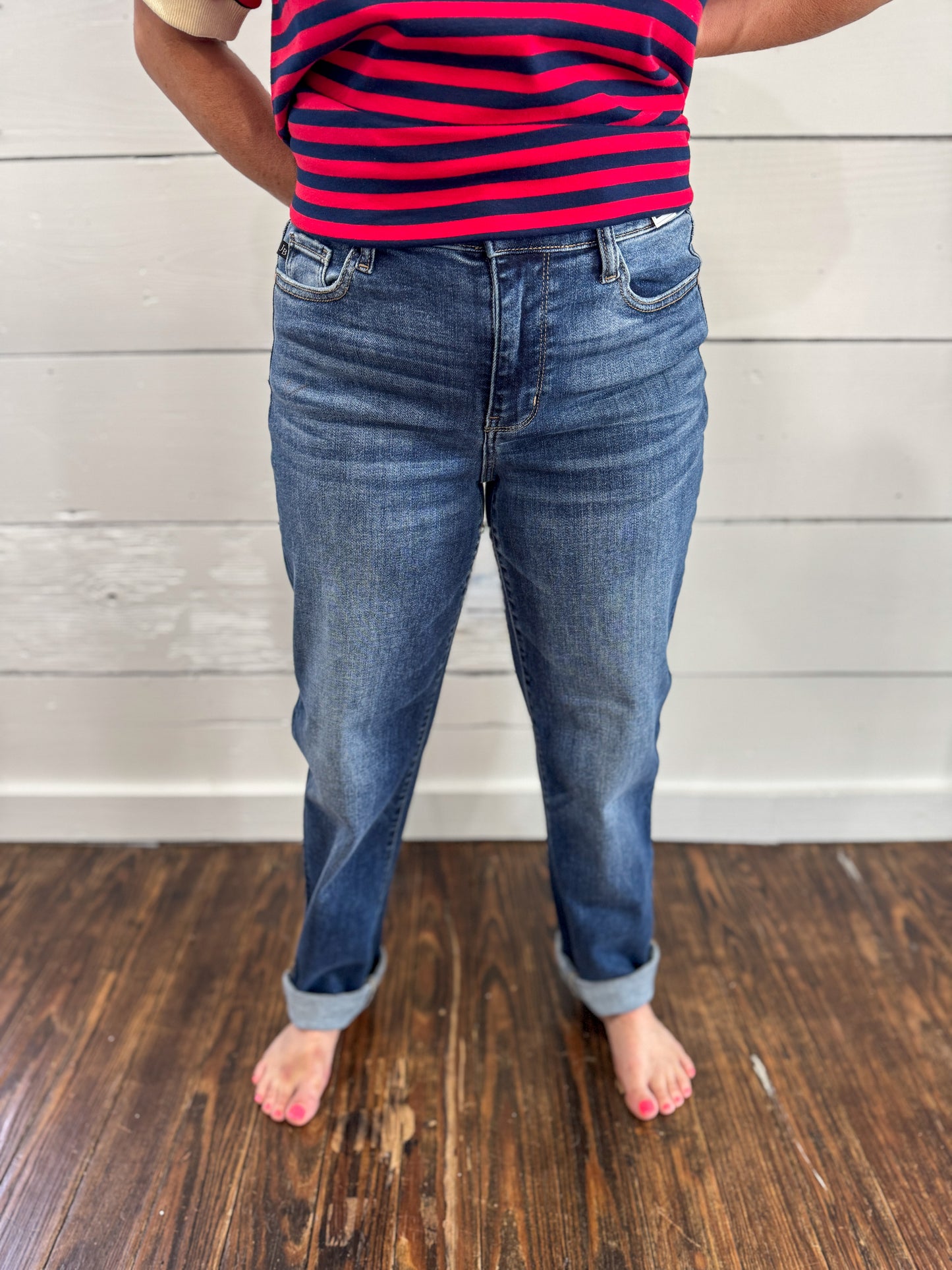 A person wearing medium-wash blue jeans with rolled cuffs, paired with a red and navy striped shirt, standing barefoot on a dark wood floor against a white shiplap wall.