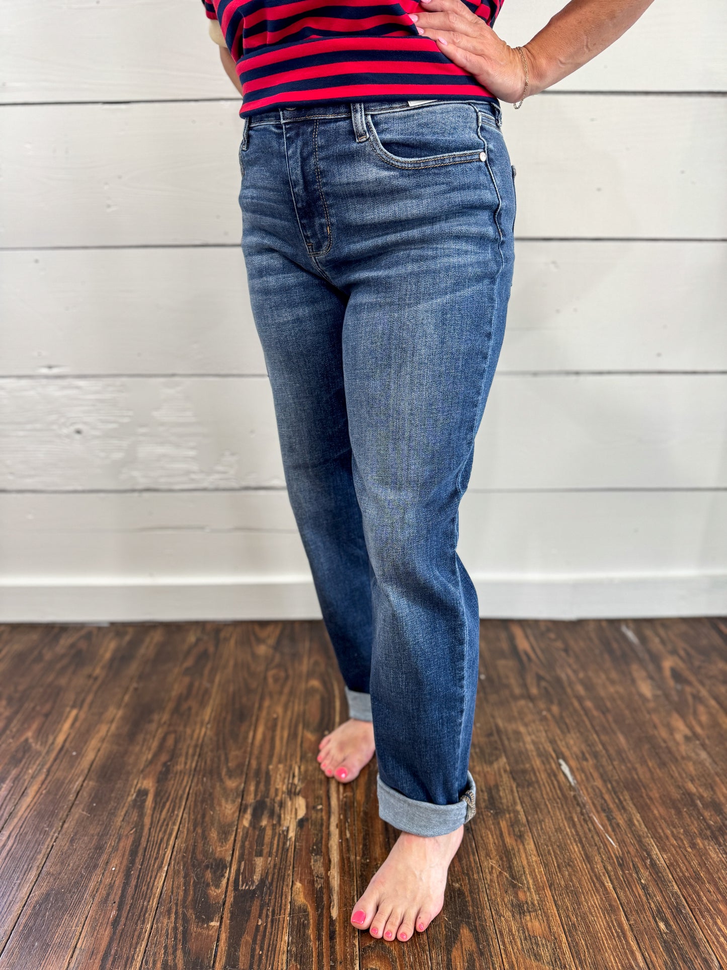 A person wearing blue denim jeans with rolled cuffs, standing barefoot on a dark wood floor against a white paneled wall. The person is also wearing a red and navy striped top, but the product focus appears to be the jeans.