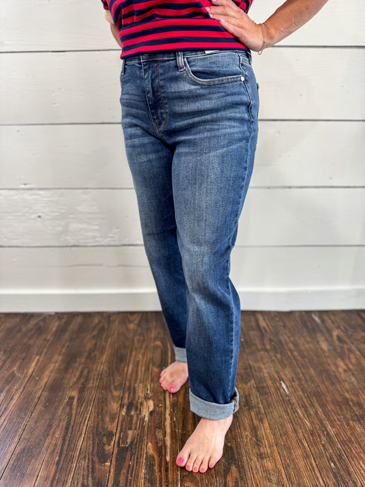 A person wearing blue denim jeans with rolled cuffs, standing barefoot on a dark wood floor against a white paneled wall. The person is also wearing a red and navy striped top, but the product focus appears to be the jeans.