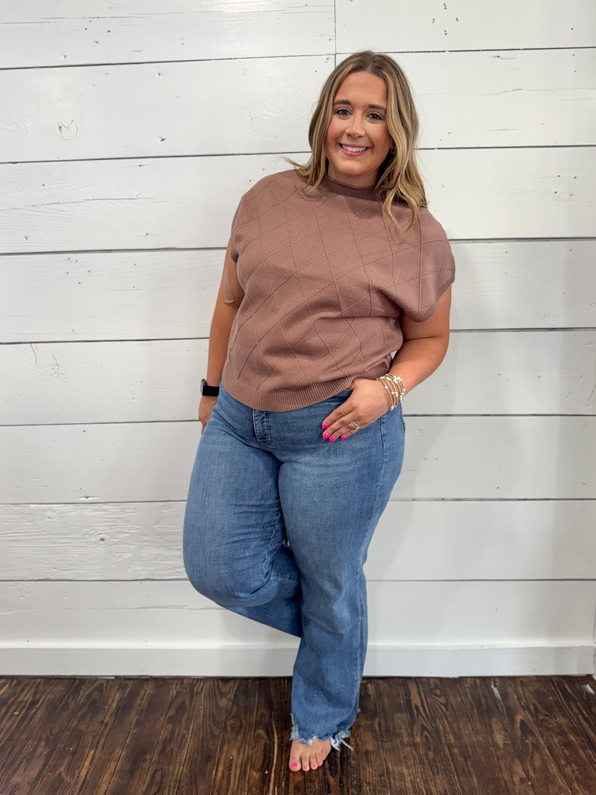 A woman modeling a short-sleeve dusty rose mock neck top and blue jeans, standing against a white shiplap wall on a dark wood floor.