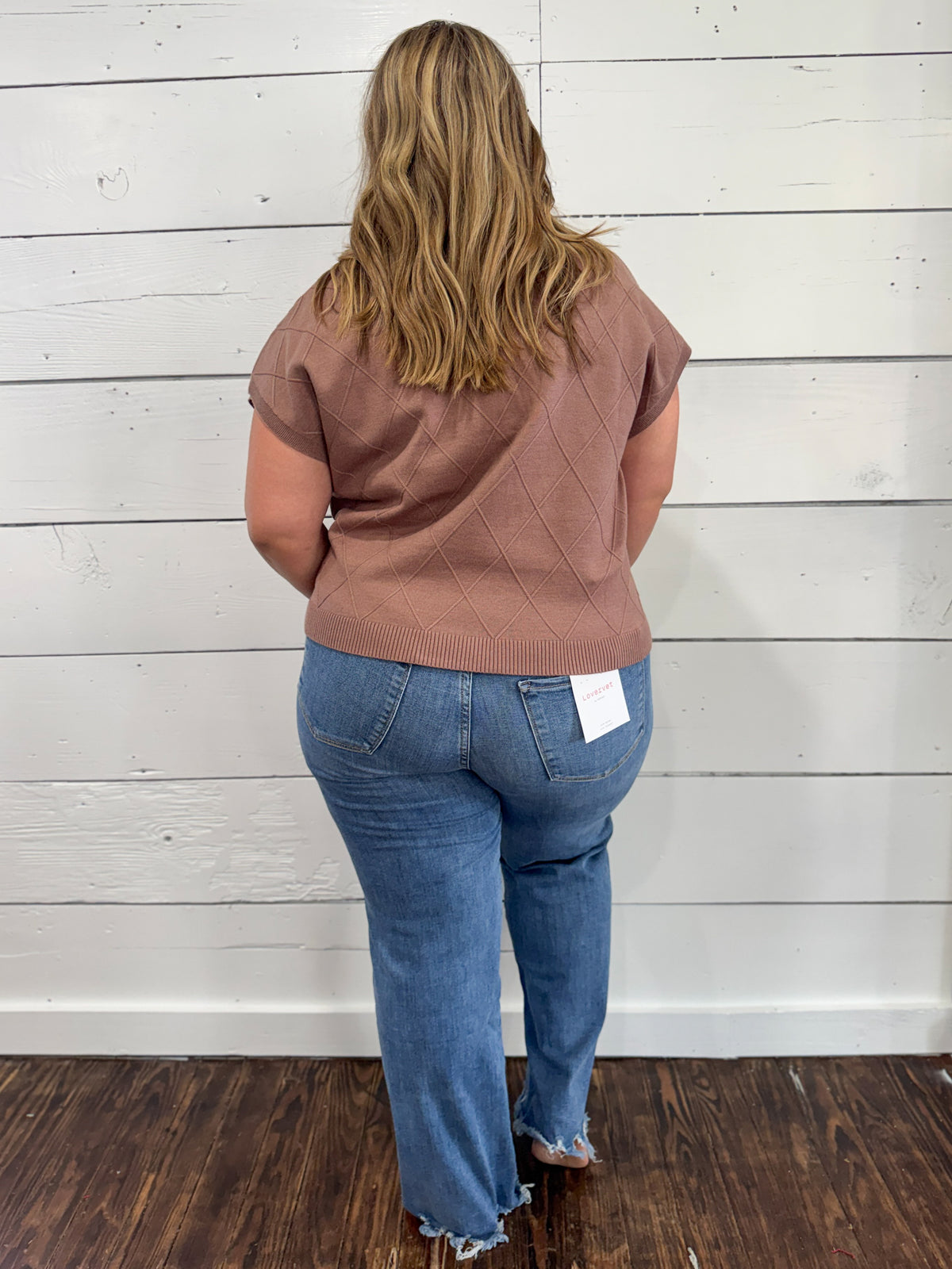 A woman viewed from behind wearing a dusty rose short-sleeve knit top and blue jeans, standing against a white wood-paneled wall on a dark wood floor.