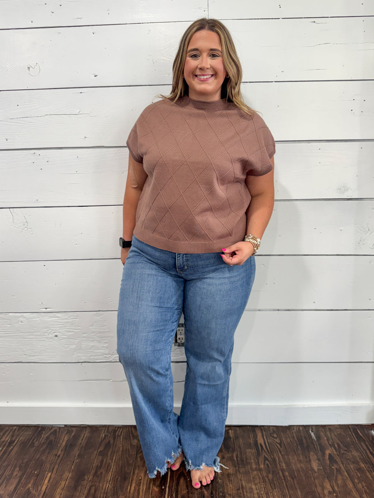 A woman modeling a short-sleeve knit top in a dusty rose-brown color with a diamond quilted pattern, paired with blue jeans, standing against a white wood-paneled wall.