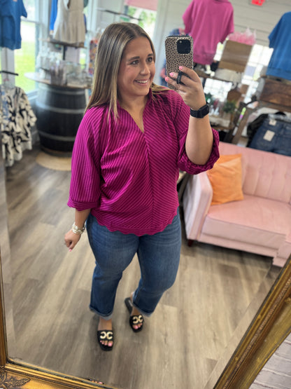 A woman takes a mirror selfie in a clothing boutique wearing a magenta pink and purple vertically striped button-up blouse, blue jeans, and black slide sandals with gold accents.