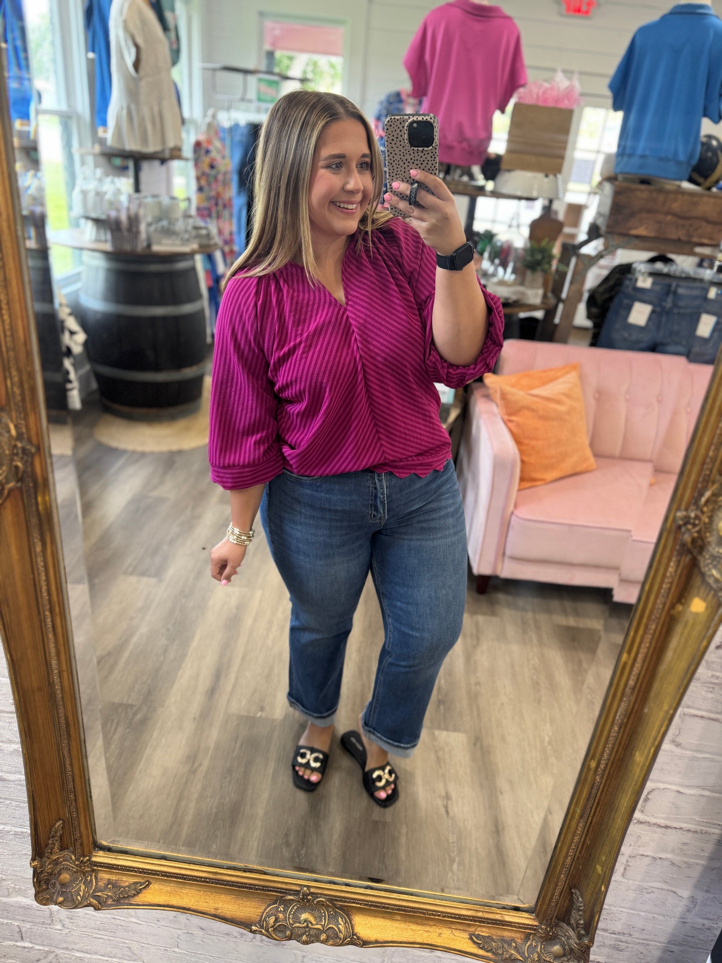 A woman takes a mirror selfie in a boutique wearing a magenta and purple vertically striped long-sleeve blouse, blue jeans, and black slide sandals with gold accents. The boutique has a large ornate gold mirror, a pink sofa with an orange pillow, and clothing racks in the background.