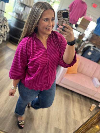A woman takes a mirror selfie in a boutique while wearing a magenta pink and purple vertically striped blouse with blue jeans.