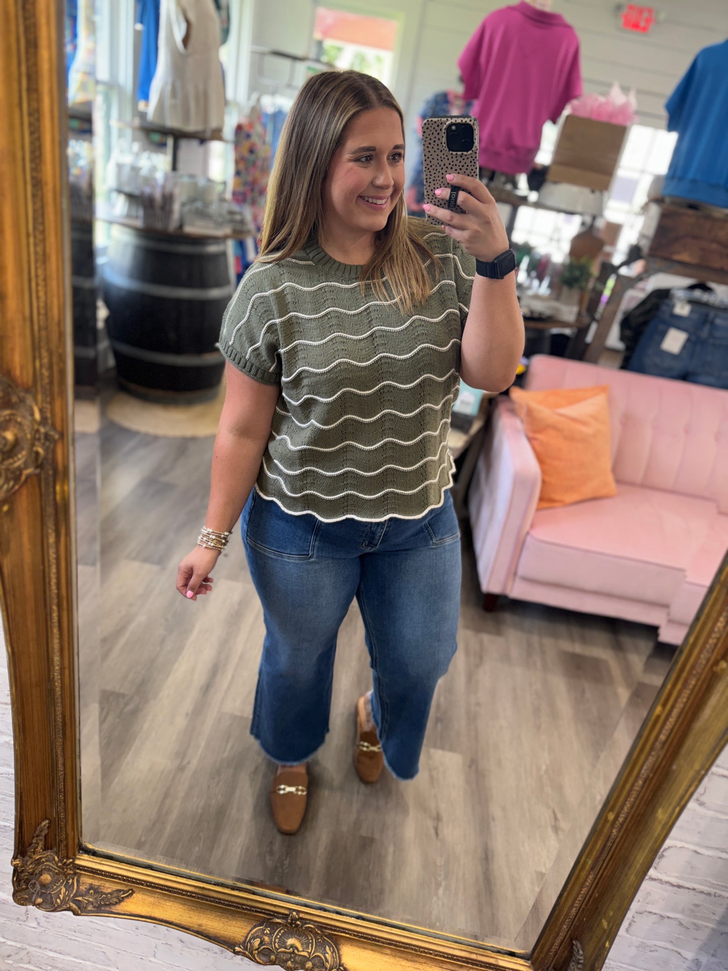 A woman takes a mirror selfie in a boutique wearing a short-sleeve olive green top with wavy horizontal stripes and blue jeans.