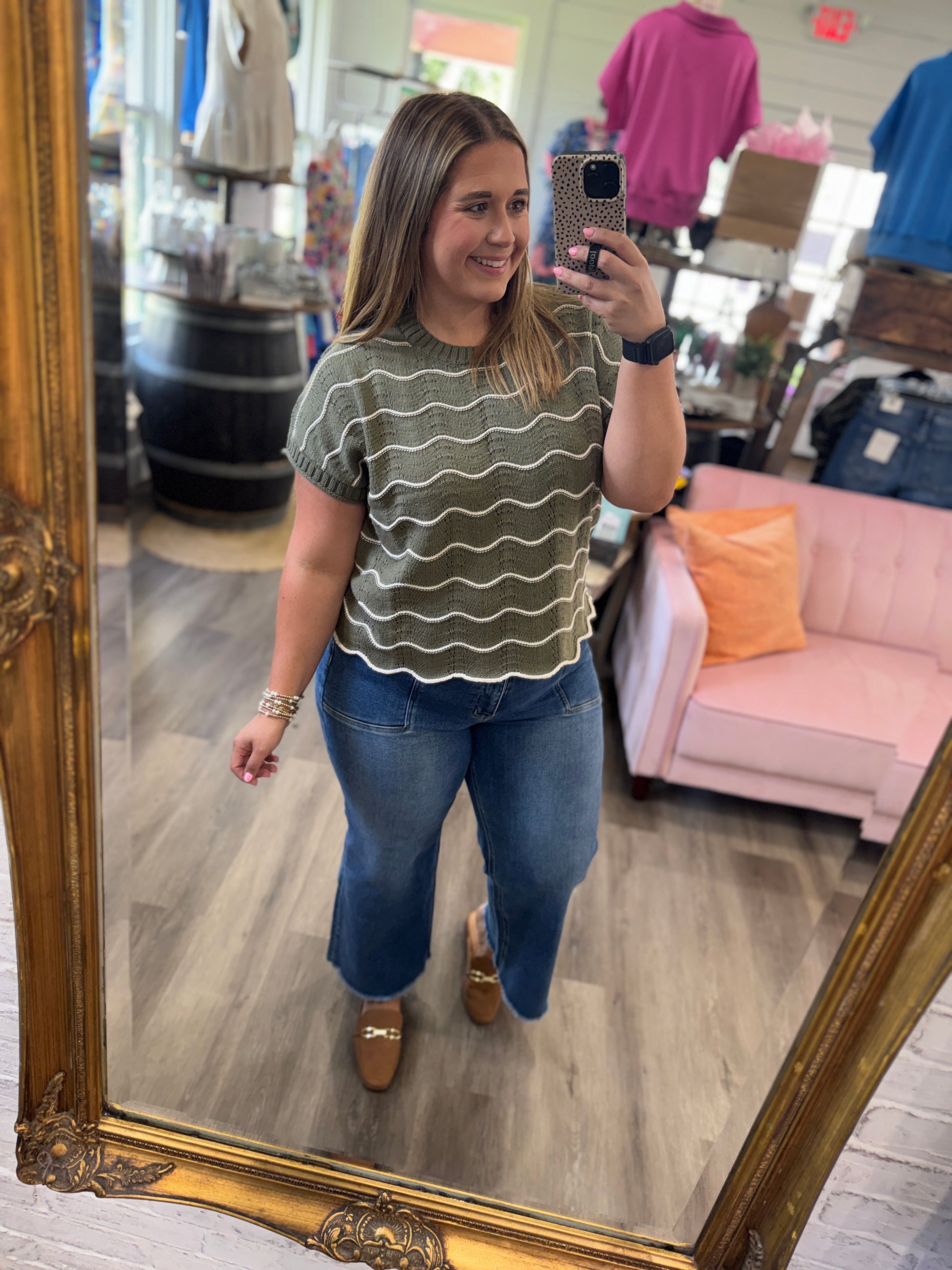 A woman takes a mirror selfie in a boutique wearing a short-sleeve olive green top with wavy horizontal stripes and blue jeans.