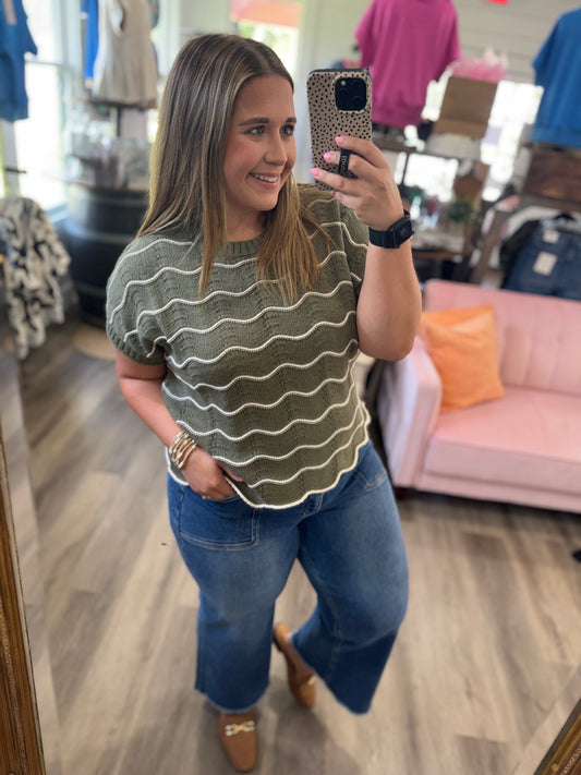 A woman taking a mirror selfie in a clothing boutique, wearing a short-sleeve olive green knit top with horizontal cream wavy stripes and blue jeans.