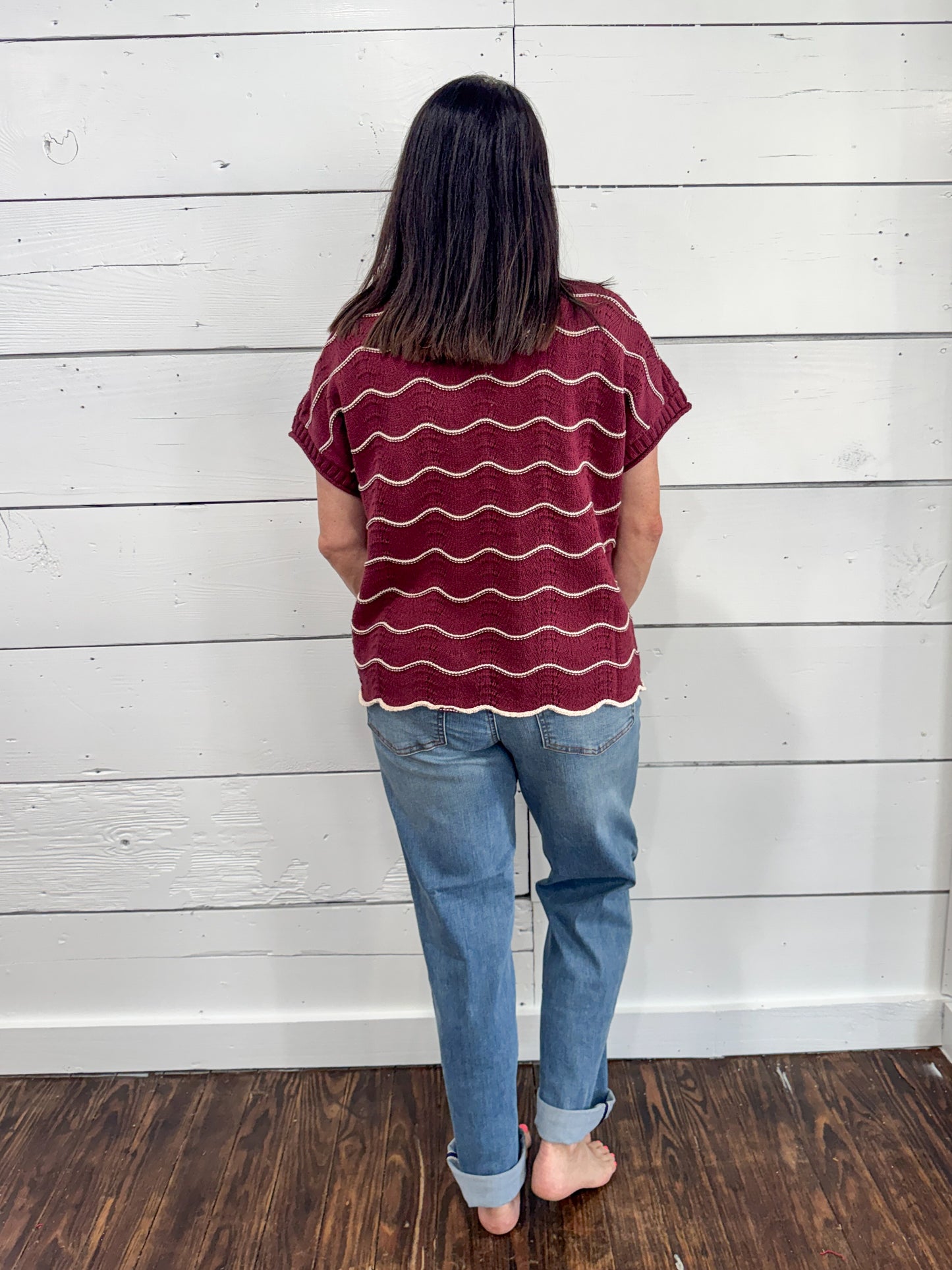 A woman viewed from behind wearing a burgundy short-sleeve t-shirt with wavy white horizontal stripes and light blue jeans, standing against a white wood-paneled wall on a dark wood floor.