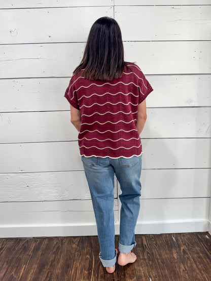 A woman viewed from behind wearing a burgundy short-sleeve t-shirt with wavy white horizontal stripes and light blue jeans, standing against a white wood-paneled wall on a dark wood floor.