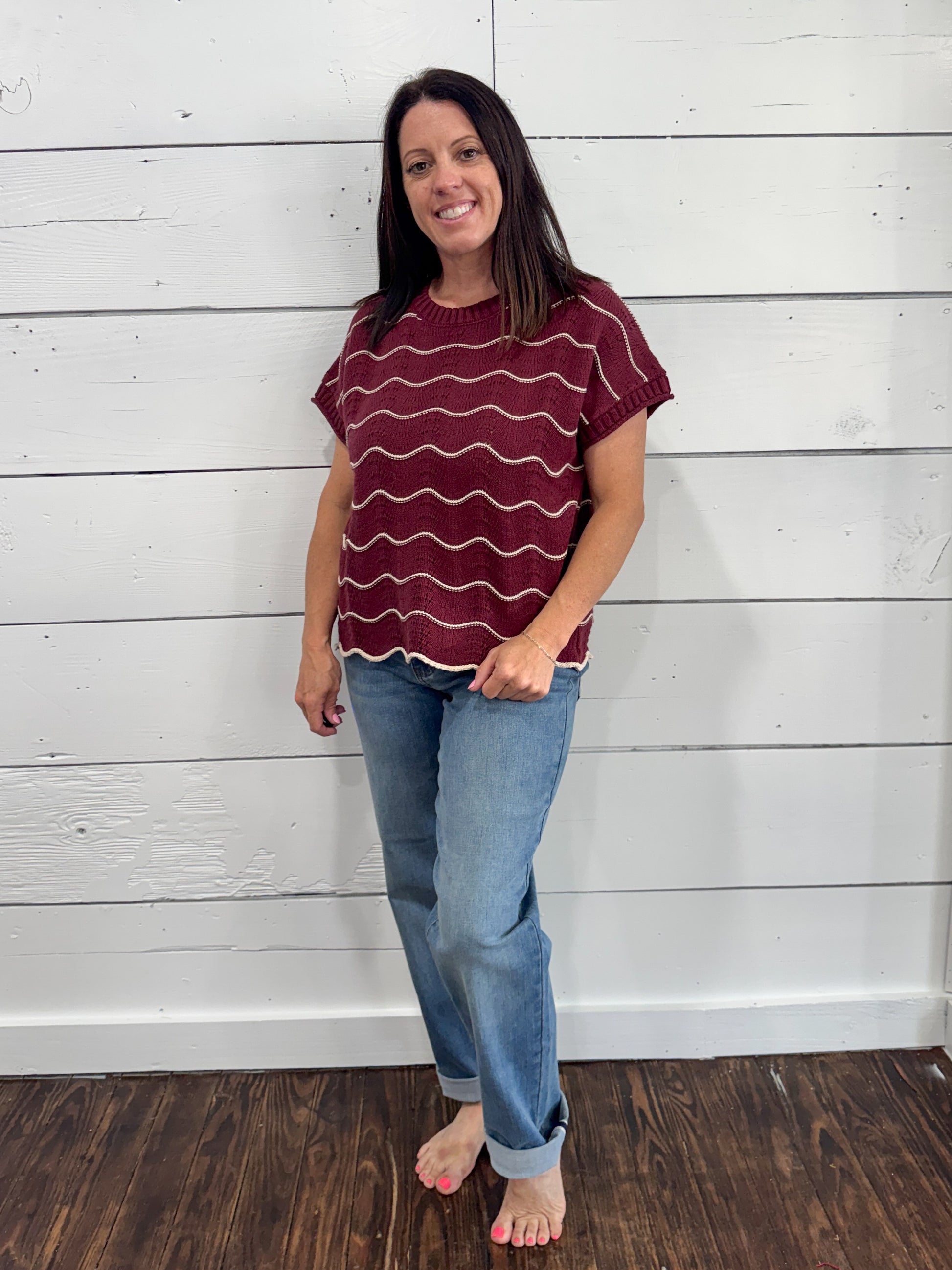 A woman stands barefoot against a white shiplap wall wearing a short-sleeve maroon top with wavy white horizontal stripes and light blue jeans.