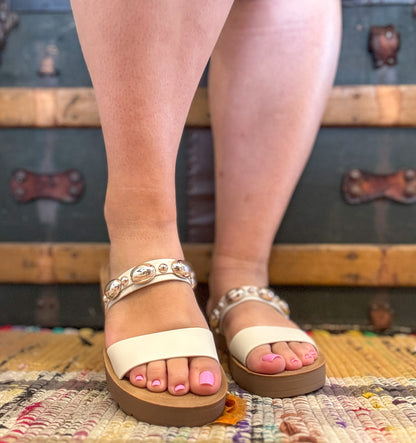 Beige sandals worn on feet with a colorful rug and wooden surface in the background