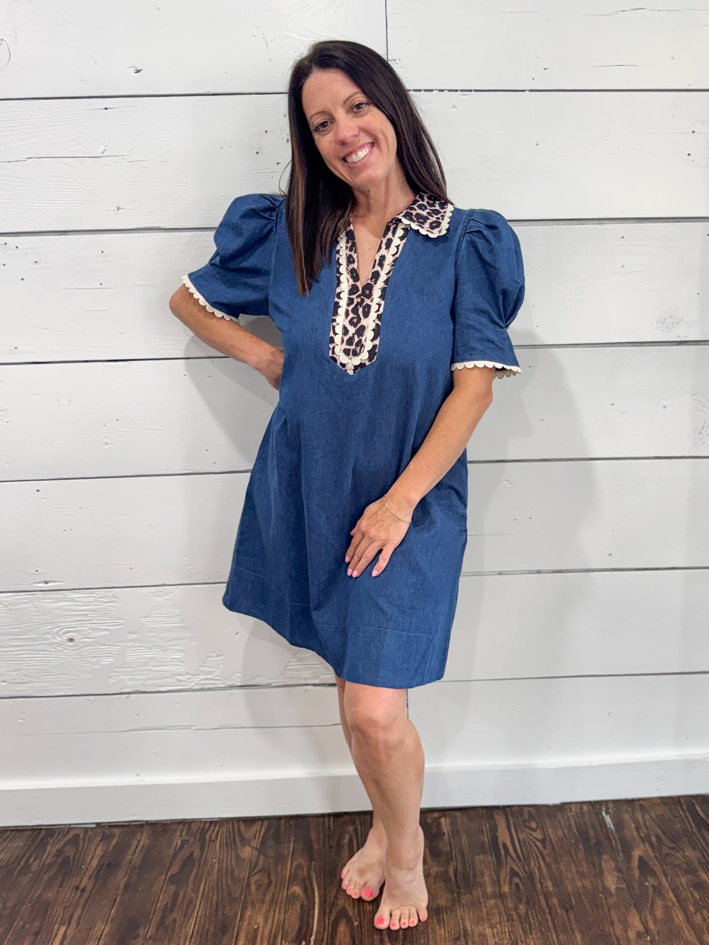 A woman modeling a short-sleeve blue dress with puff sleeves and a decorative beige and black patterned collar, standing against a white shiplap wall.