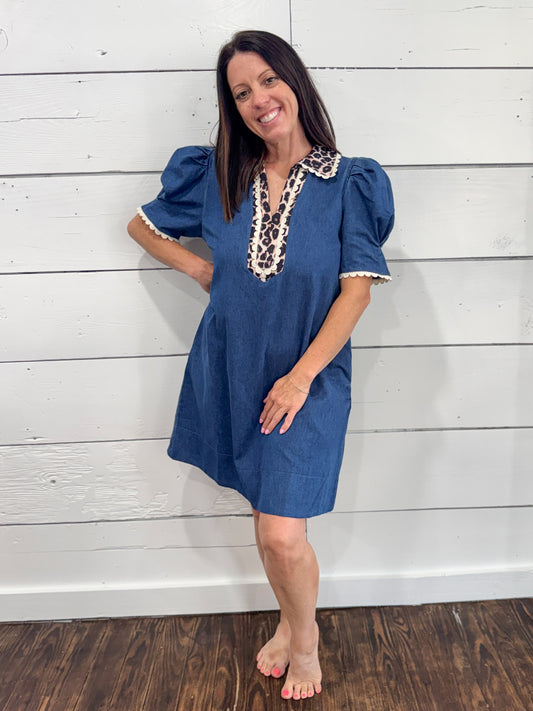 A woman modeling a short-sleeve blue dress with puff sleeves and a decorative beige and black patterned collar, standing against a white shiplap wall.