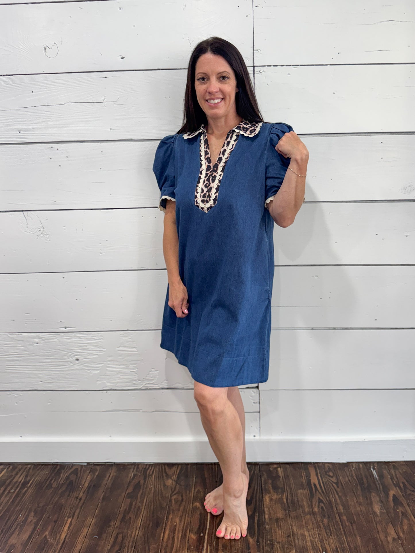 A woman modeling a blue short-sleeve tunic dress with a decorative cream lace neckline, standing against a white shiplap wall and dark wood floor.