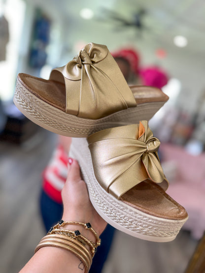 Gold sandals with bow detail held by a person in a store setting