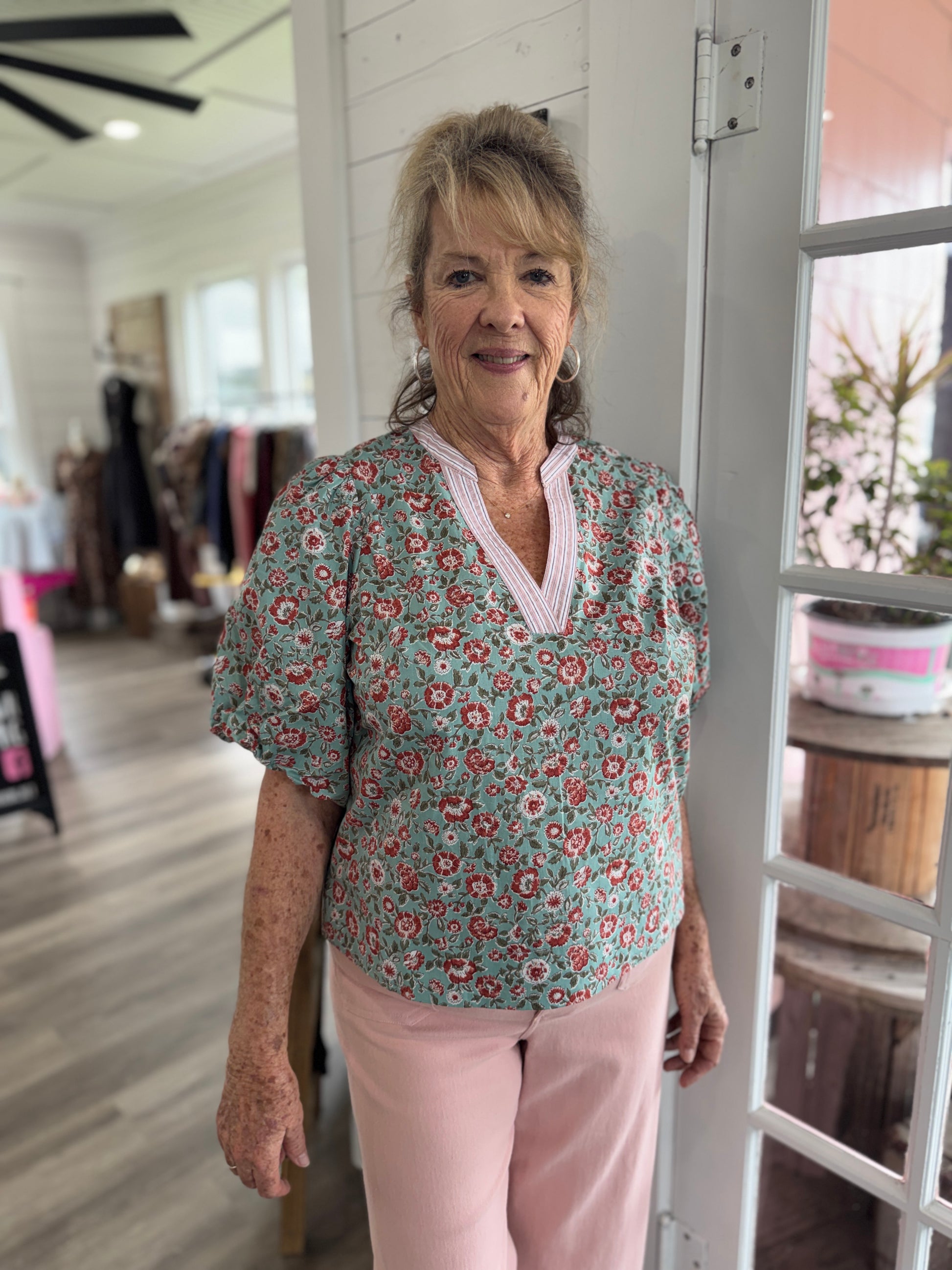 Woman wearing a floral blouse and pink pants standing indoors.