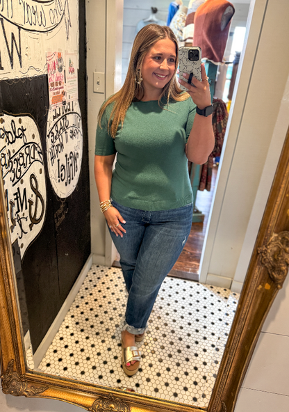 Woman taking a mirror selfie wearing a green top and blue jeans in a store.