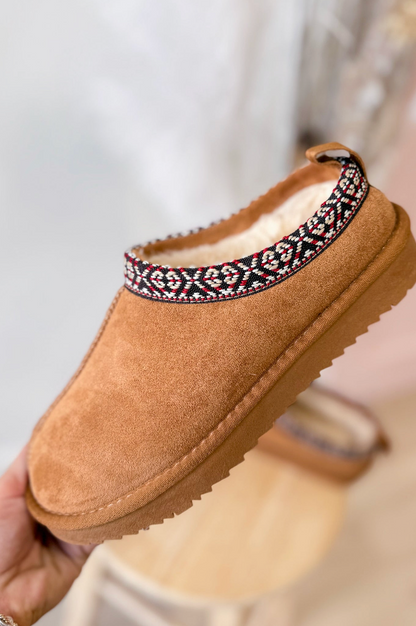 Brown slipper with decorative band held by a hand against a neutral background
