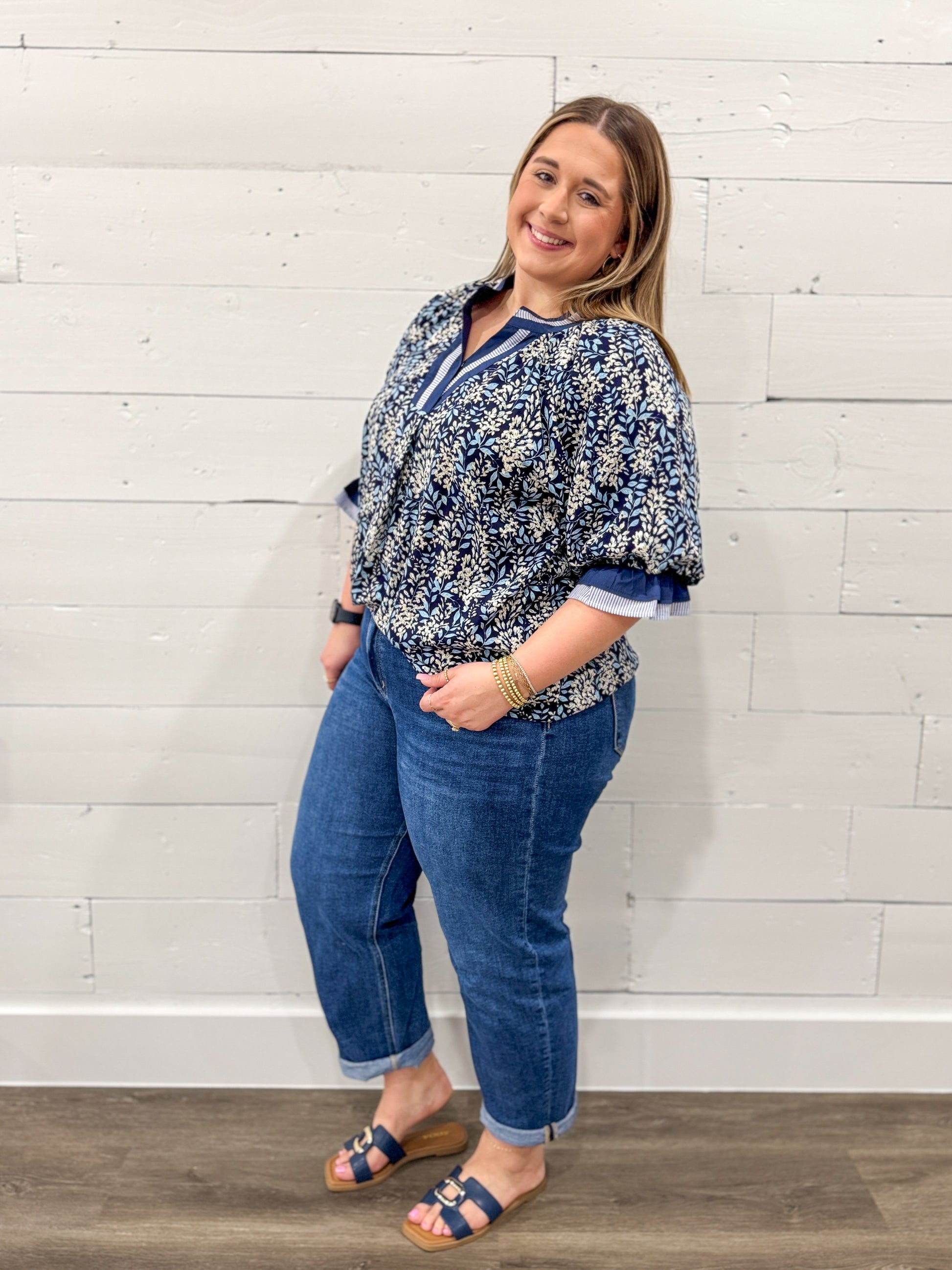 Woman wearing a blue floral blouse and jeans standing against a white brick wall.